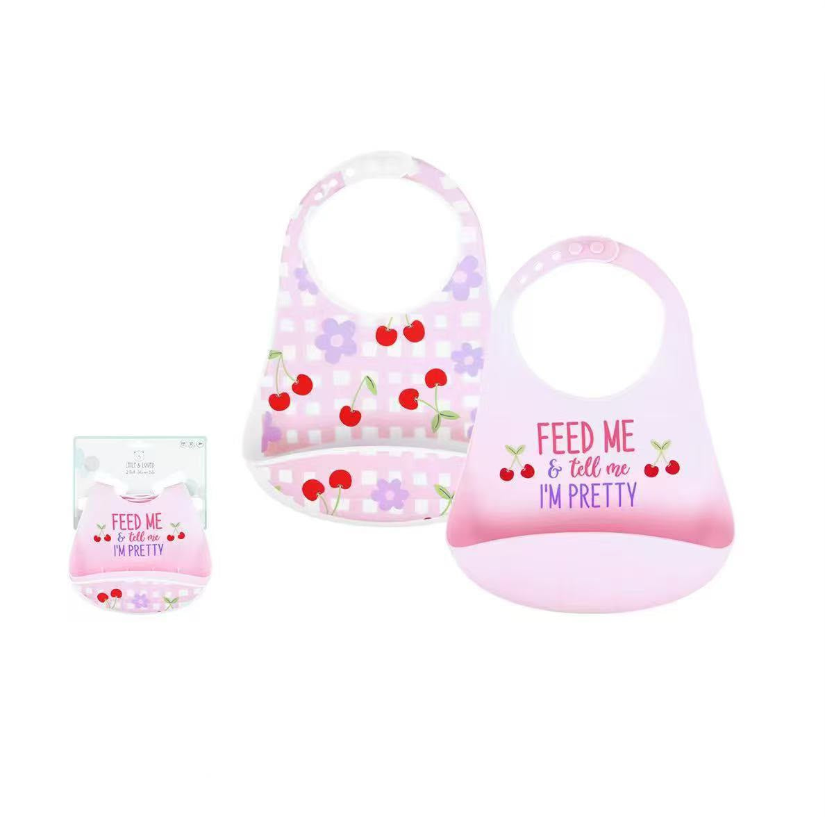 Two silicone baby bibs shown on a white background with product text and a small packaged view. The left bib is light lavender with white polka dots and red cherry prints; the right bib is pale pink with a gradient and the phrase 'FEED ME & tell me I'M PRETTY' plus cherry prints.