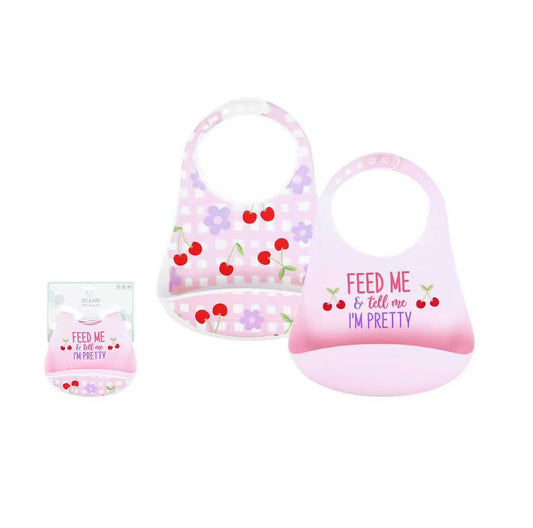 Two silicone baby bibs shown on a white background with product text and a small packaged view. The left bib is light lavender with white polka dots and red cherry prints; the right bib is pale pink with a gradient and the phrase 'FEED ME & tell me I'M PRETTY' plus cherry prints.
