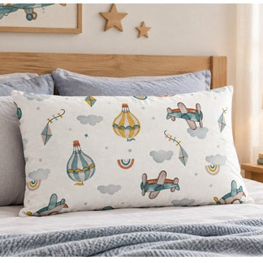 A white pillow sham with a colorful children's airplane and hot air balloon pattern, styled on a bed with gray pillows and a wooden headboard in a bright bedroom.