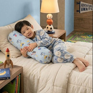 A young boy wearing blue pajamas lies on a bed using a light blue contoured lumbar pillow with cartoon animal prints. The bedroom setting includes a wooden nightstand with a lamp and rocket toy, and a blue wall in the background.